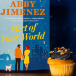 The book 'Part of Your World' by Abby Jimenez stands upright against a deep blue background with starry accents. The cover features an illustration of a man and woman holding hands, with a small white dog beside them, set against a city skyline at night. To the right of the book is a yellow cupcake with dark blue frosting, decorated with gold and white star-shaped sprinkles, adding a whimsical touch to the scene.